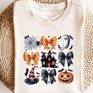 May include: A cream-coloured t-shirt with a Halloween design. The design includes a spiderweb with a spider, orange bows, a haunted house, a ghost, a witch's hat, black bows, and a jack-o'-lantern. The shirt is displayed on a woven surface.