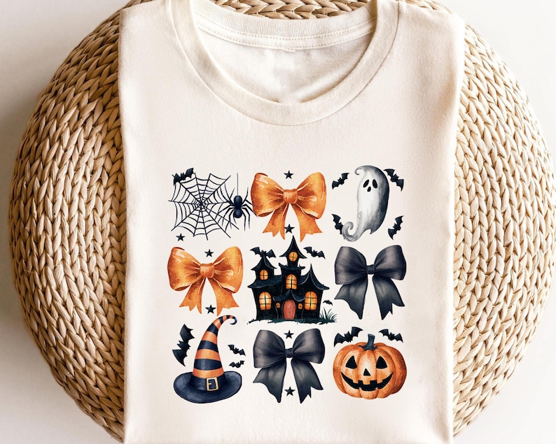 May include: A cream-coloured t-shirt with a Halloween design. The design includes a spiderweb with a spider, orange bows, a haunted house, a ghost, a witch's hat, black bows, and a jack-o'-lantern. The shirt is displayed on a woven surface.