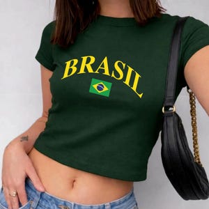 May include: A dark green short-sleeved crop top featuring the word BRASIL printed in a yellow arched font above a small rectangular graphic of the Brazilian flag. The garment has a crew neckline and a fitted silhouette.