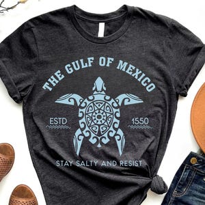 May include: Dark gray t-shirt with a light blue graphic of a sea turtle and the text "THE GULF OF MEXICO" and "ESTD 1550 STAY SALTY AND RESIST". The shirt is folded and knotted at the bottom.