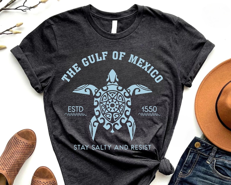 May include: Dark gray t-shirt with a light blue graphic of a sea turtle and the text "THE GULF OF MEXICO" and "ESTD 1550 STAY SALTY AND RESIST". The shirt is folded and knotted at the bottom.