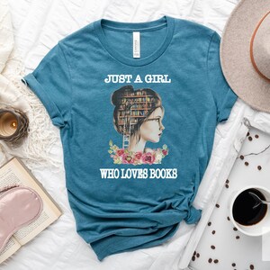 Pu&ograve; includere: Maglietta blu turchese con una grafica di una donna con una pila di libri come testa. Il testo "Just a girl who loves books" &egrave; stampato sulla maglietta.