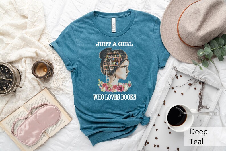 Pu&ograve; includere: Maglietta blu turchese con una grafica di una donna con una pila di libri come testa. Il testo "Just a girl who loves books" &egrave; stampato sulla maglietta.