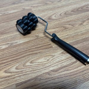 May include: A black massage roller tool featuring a cylindrical head with multiple rounded protrusions attached to a silver metal frame and a black textured handle. The device is designed for manual use, with the roller head rotating on the frame.