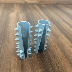 Op de afbeelding: Twee lichtblauwe cilindrische voorwerpen van plastic met een rij spikes rond de boven- en onderkant. De voorwerpen liggen op een houten oppervlak.