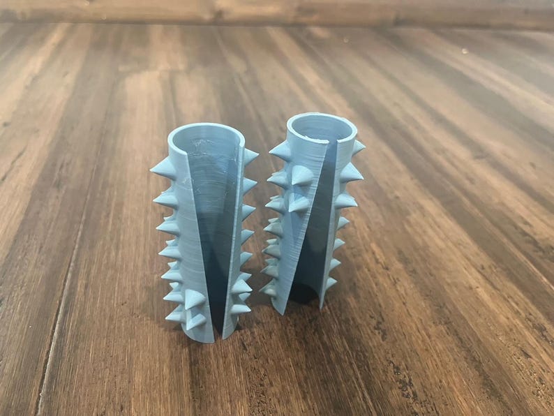 Op de afbeelding: Twee lichtblauwe cilindrische voorwerpen van plastic met een rij spikes rond de boven- en onderkant. De voorwerpen liggen op een houten oppervlak.