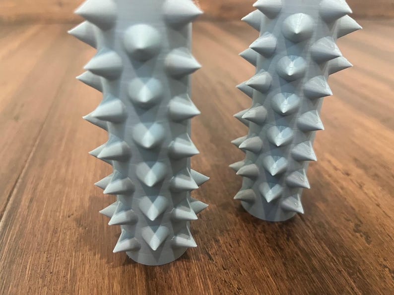 Op de afbeelding: Twee lichtblauwe, stekelige, cilindrische voorwerpen. De voorwerpen zijn gemaakt van een zacht, rubberachtig materiaal en hebben een getextureerd oppervlak. De voorwerpen zijn vergelijkbaar in grootte en vorm.