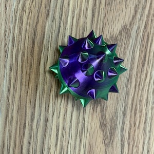 Könnte beinhalten: Ein kugelförmiges Objekt mit einer metallischen Oberfläche, die zwischen Violett und Grün wechselt. Die Oberfläche ist mit zahlreichen spitzen, kegelförmigen Dornen in einem radialen Muster bedeckt, was eine texturierte Optik erzeugt.