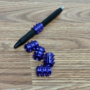 Op de afbeelding: Een zwarte balpen met een getextureerde grip en een paarse, stekelige, cilindrische pen-topper, vergezeld van vier extra bijpassende paarse, stekelige, cilindrische pen-toppers die in een klein groepje bij elkaar liggen.
