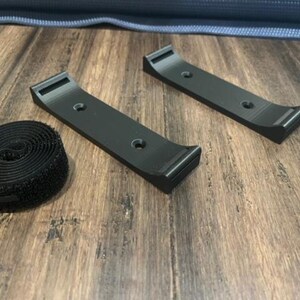 May include: Two black 3D-printed brackets and a roll of black hook-and-loop fastener tape. The brackets have a curved design with mounting holes. The items are on a wood-grain surface.