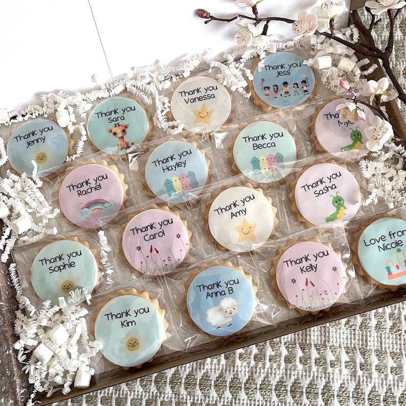 May include: A collection of round, scalloped-edge biscuits individually wrapped in clear plastic, each featuring a unique printed design and a name such as Jenny, Sara, Vanessa, Jess, Becca, Rachel, Hayley, Amy, Sasha, Carol, Sophie, Anna B, Kelly, and Nicole.