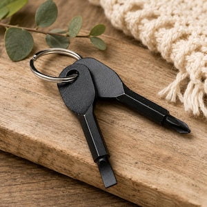 May include: A set of two black multi-tool screwdrivers attached to a silver key ring. One screwdriver has a flathead tip, and the other has a Phillips head tip. The tools are on a wooden surface with a neutral-colored knitted item in the background.