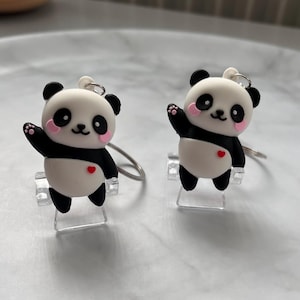 May include: A pair of panda bear earrings with black and white fur, pink cheeks, and a red heart on their chest. The earrings are attached to silver hoops.