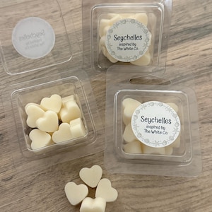 May include: Several clear plastic containers filled with heart-shaped wax melts. Some containers have a circular label that reads "Seychelles inspired by The White Co." The wax melts are a creamy off-white color.