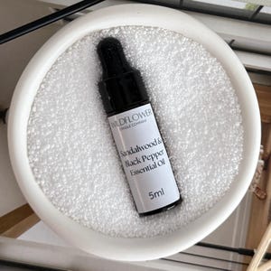 Puede incluir: Una peque&ntilde;a botella negra de aceite esencial de s&aacute;ndalo y pimienta negra de Wildflower Candle Company. La botella contiene 5 ml de aceite y est&aacute; sobre una superficie blanca.