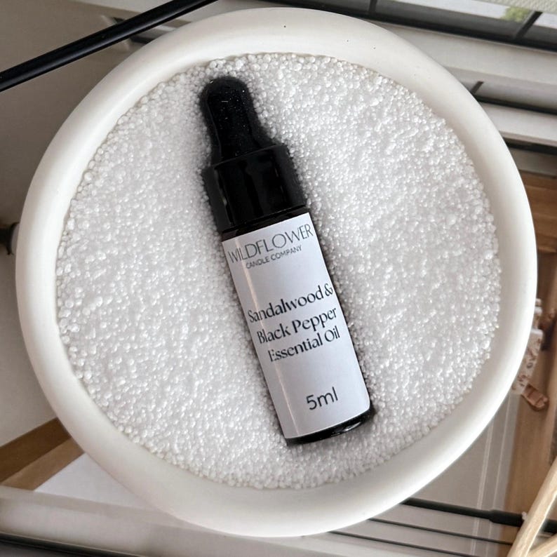 Puede incluir: Una peque&ntilde;a botella negra de aceite esencial de s&aacute;ndalo y pimienta negra de Wildflower Candle Company. La botella contiene 5 ml de aceite y est&aacute; sobre una superficie blanca.