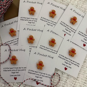 Peut inclure: Un ensemble de huit petites cartes blanches avec un autocollant en pain d'épices sur chacune. Les cartes portent le texte "A Pocket Hug" et un message sur l'amour et les câlins.
