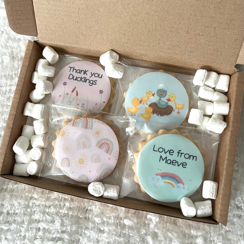 May include: A cardboard box containing four round biscuits individually wrapped in clear plastic, surrounded by white mini marshmallows. The biscuits feature printed designs including ducks, rainbows, suns, and the text Thank you Ducklings and Love from Maeve.