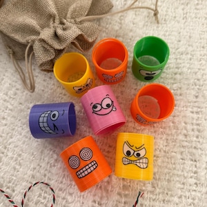 Puede incluir: Colección de juguetes de resorte de plástico de colores, cada uno con una cara de dibujos animados diferente. Los resortes son de varios colores, incluyendo naranja, amarillo, rosa, verde y morado. Una pequeña bolsa de yute está en el fondo.