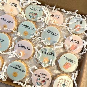May include: A collection of round, individually wrapped biscuits arranged in a cardboard box with white paper shred. Each biscuit features a unique name or phrase like Alfie, Esme, Holly, Ellie, Maryam, Sienna, James, Ana, Liliana, Tamsin, Maddox, Mrs Shuttleworth, and We're proud of you, accompanied by colourful illustrations of suns, stars, and school supplies.