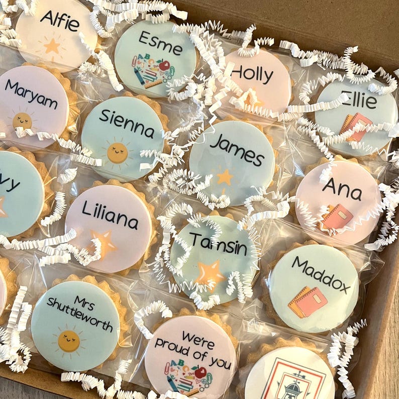 May include: A collection of round, individually wrapped biscuits arranged in a cardboard box with white paper shred. Each biscuit features a unique name or phrase like Alfie, Esme, Holly, Ellie, Maryam, Sienna, James, Ana, Liliana, Tamsin, Maddox, Mrs Shuttleworth, and We're proud of you, accompanied by colourful illustrations of suns, stars, and school supplies.