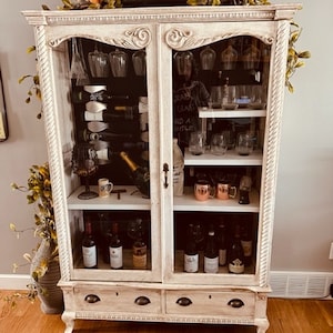 May include: White wooden cabinet with glass doors and two drawers. The cabinet is filled with wine bottles, glasses, and other barware. The cabinet has a distressed white finish and ornate details.