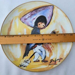 K&ouml;nnte beinhalten: Dekorative Platte mit einer skurrilen Illustration eines Kindes mit einem Besen, das eine blaue Jacke und eine wei&szlig;e Hose tr&auml;gt. Die Platte hat einen Goldrand und die Signatur des K&uuml;nstlers, De Grazia, mit "Arizona USA" darunter. Die Platte misst etwa 25 cm im Durchmesser.