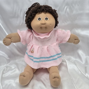 Pode incluir: Boneca vintage com cabelo castanho e tom de pele claro. A boneca usa um vestido de malha rosa com listras azuis e um pequeno emblema de pinguim. A boneca está sentada com os braços estendidos.