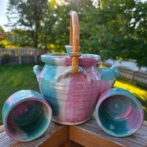 May include: A handmade ceramic teapot and two matching cups. The teapot has a wooden handle and is painted with vertical stripes of turquoise, white, and pink. The cups have a similar color scheme. The set is displayed on a wooden surface.
