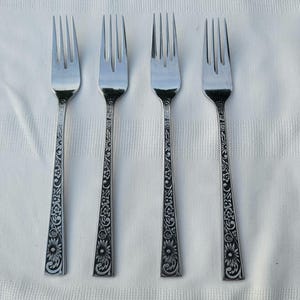 May include: Four silver forks with ornate floral and scrollwork designs on the handles. The forks are arranged on a white surface, showcasing their polished appearance and intricate detailing. The tines are evenly spaced.