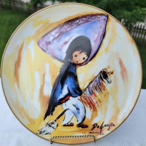 K&ouml;nnte beinhalten: Dekorative Platte mit Goldrand, die eine stilisierte Illustration einer Figur mit einem Pferd zeigt. Die Figur tr&auml;gt einen blauen Mantel und eine wei&szlig;e Hose und h&auml;lt einen Besen. Die Platte hat einen gelb-orangen Hintergrund mit der Signatur des K&uuml;nstlers.