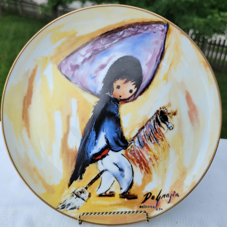 K&ouml;nnte beinhalten: Dekorative Platte mit Goldrand, die eine stilisierte Illustration einer Figur mit einem Pferd zeigt. Die Figur tr&auml;gt einen blauen Mantel und eine wei&szlig;e Hose und h&auml;lt einen Besen. Die Platte hat einen gelb-orangen Hintergrund mit der Signatur des K&uuml;nstlers.