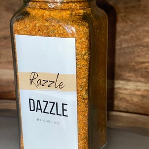 May include: A clear glass jar with a silver lid filled with a blend of spices. The label reads "Razzle Dazzle by Chef Nic" in black text. The spices are a mix of orange, brown, and green.