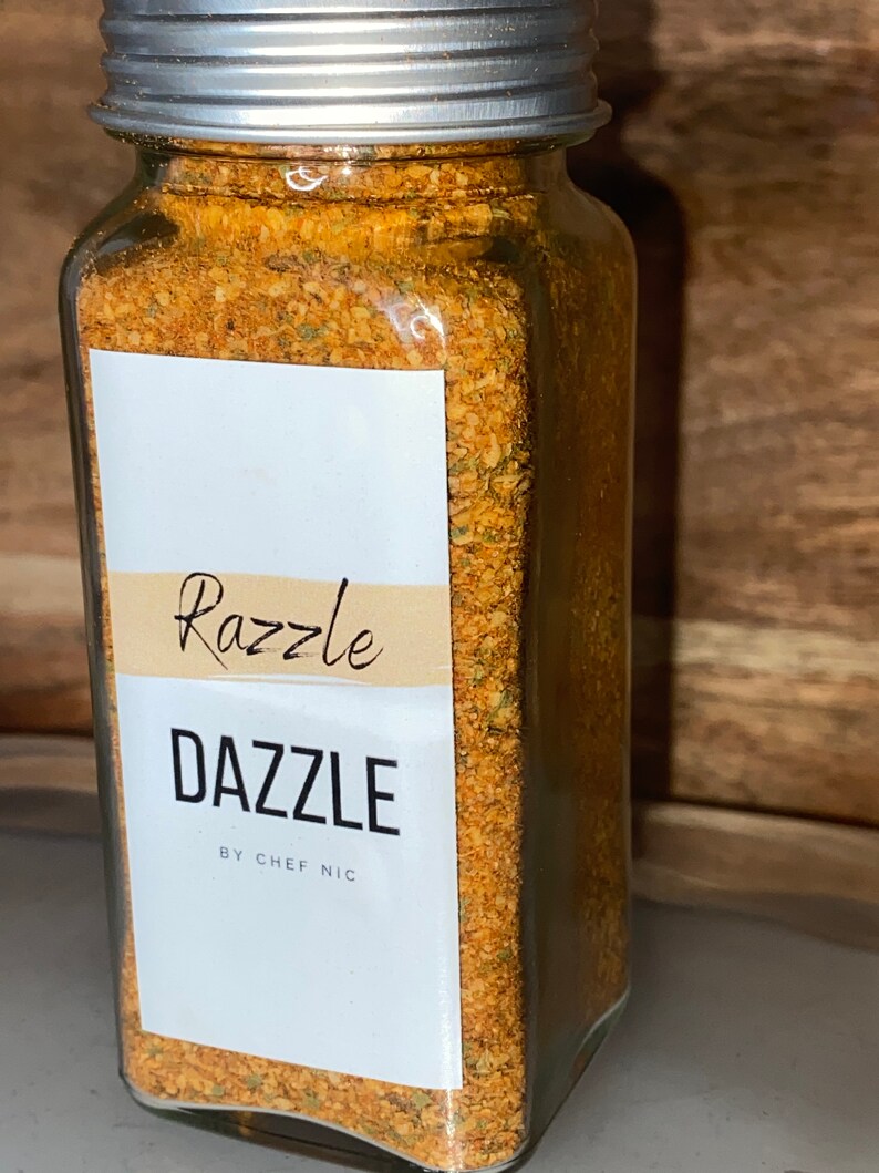 May include: A clear glass jar with a silver lid filled with a blend of spices. The label reads "Razzle Dazzle by Chef Nic" in black text. The spices are a mix of orange, brown, and green.