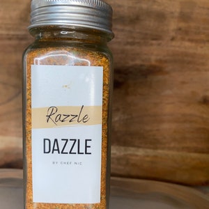 May include: A clear glass jar with a silver lid filled with a yellow-orange spice blend. The jar has a white label with the words "Razzle Dazzle" and "BY CHEF NIC" in black text. The background is a wooden surface.