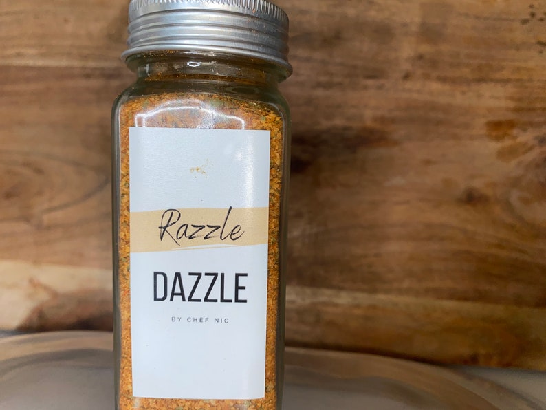 May include: A clear glass jar with a silver lid filled with a yellow-orange spice blend. The jar has a white label with the words "Razzle Dazzle" and "BY CHEF NIC" in black text. The background is a wooden surface.