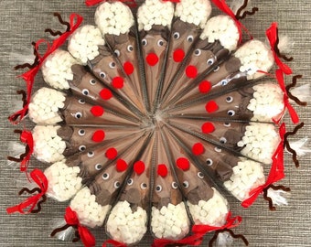 May include: A circular arrangement of cone-shaped cellophane bags filled with hot chocolate mix and marshmallows. Each bag is decorated to resemble a reindeer, with googly eyes, a red nose, and brown pipe cleaner antlers. Red ribbons secure the bags.