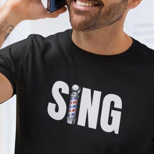Puede incluir: Una camiseta negra con la palabra "SING" impresa en letras blancas. Un poste de barbero está incorporado en la "I" de la palabra "SING".