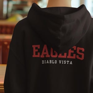May include: Black hooded sweatshirt with the word "EAGLES" in red block letters and "DIABLO VISTA" in white, centered on the back. The sweatshirt is worn by a person with red hair, viewed from the back.