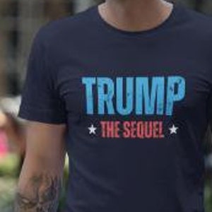 Może przedstawiać: Koszulka w kolorze granatowym z napisem "TRUMP *THE SEQUEL*" w niebieskich i różowych literach.