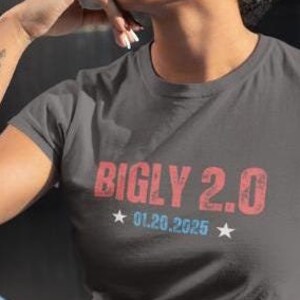 Może przedstawiać: Szary t-shirt z nadrukiem "BIGLY 2.0" w czerwono-niebieskich literach. Data "01.20.2025" jest wydrukowana pod tekstem na biało.