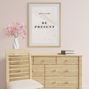 May include: A framed print with a light pink background and the text "note to self: BE PRESENT" in black. The print is hanging on a light pink wall above a wooden dresser with six drawers. A vase with pink flowers is on the left side of the print.