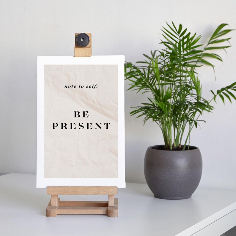 May include: A white canvas with the text "note to self: BE PRESENT" on a beige marble background. The canvas is on a wooden easel and a potted plant is in the background.
