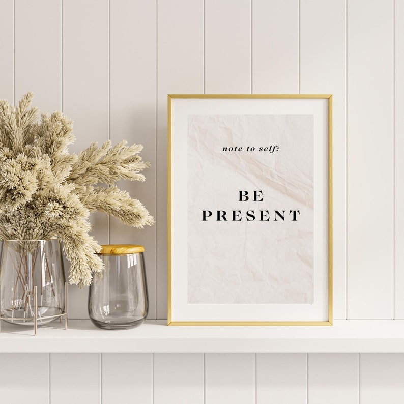 May include: A gold framed print with a white background and black text that reads "note to self: BE PRESENT". The print is sitting on a white shelf with a vase of dried flowers to the left.