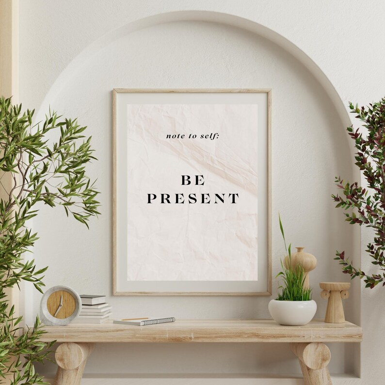 May include: A framed print with a beige background and the text "note to self: BE PRESENT" in black lettering. The print is hanging on a white wall with a wooden frame.