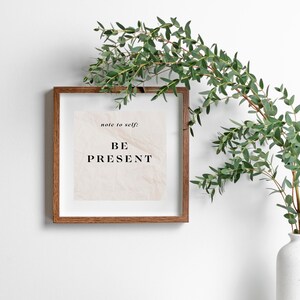 May include: A framed print with a white background and the text "note to self: BE PRESENT" in black lettering. The frame is brown wood and is hanging on a white wall. A sprig of green eucalyptus is in the background.