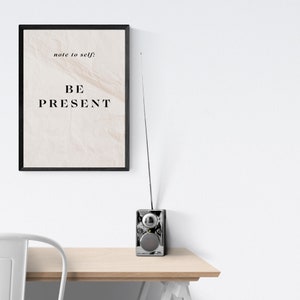 May include: A black framed print with the text "note to self: BE PRESENT" on a white background. A silver radio with a long antenna is on a wooden table.