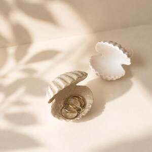 Könnte beinhalten: Zwei muschelförmige Schmuckschalen. Eine ist geöffnet und enthält goldene Ringe. Die andere ist leer. Beide sind cremefarben mit strukturierten Oberflächen. Der Hintergrund ist ein neutrales Beige mit Blätterschatten.