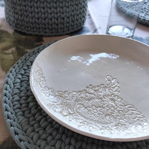 Può includere: Un piatto in ceramica bianca con un motivo floreale poggia su un sottobicchiere all'uncinetto grigio. Un cestino all'uncinetto abbinato contiene tovaglioli. Un bicchiere trasparente è anche sul tavolo. L'apparecchiatura ha un'estetica rustica fatta a mano.