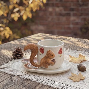 Könnte beinhalten: Ein Set aus weißer Keramiktasse und Untertasse. Die Tasse hat einen dreidimensionalen braunen Eichhörnchenhenkel und bemalte rote Pilzmotive. Die Untertasse ist blattförmig und die Tasse besitzt einen gewellten, unebenen Rand mit Textur.